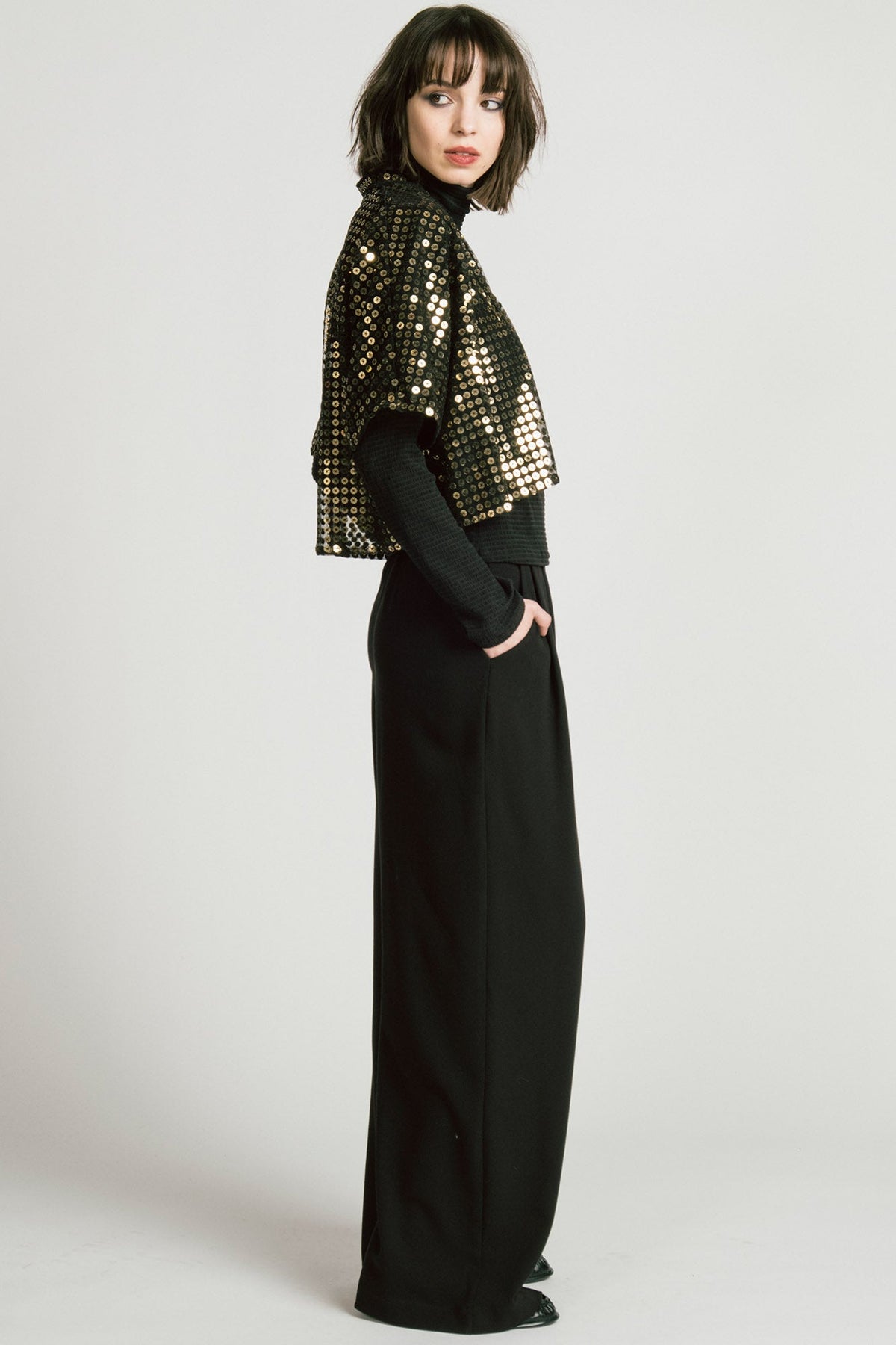 Side view of a woman wearing the Corvus Shrug by Allison Wonderland in Sequins, a sheer black bolero with golden sequins. She is wearing it over a black turtleneck and black pants and standing in front of a white background. 