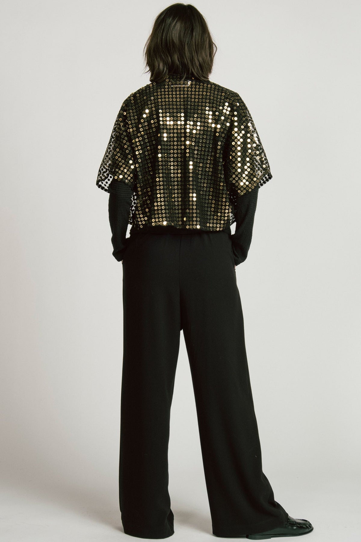 Back view of a woman wearing the Corvus Shrug by Allison Wonderland in Sequins, a sheer black bolero with golden sequins. She is wearing it over a black turtleneck and black pants and standing in front of a white background. 