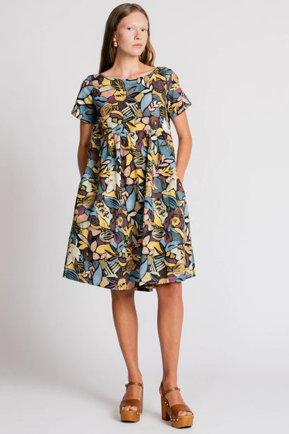 A woman wearing the Lark Dress by Allison Wonderland in Black Floral print, a short sleeved babydoll dress with pockets and a crossover back. She is standing in front of a plain background. 