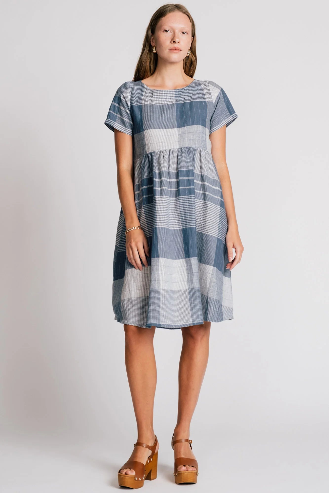 A woman wearing the Lark Dress by Allison Wonderland in Blue Plaid print, a short sleeved babydoll dress with pockets and a crossover back. She is standing in front of a plain background. 