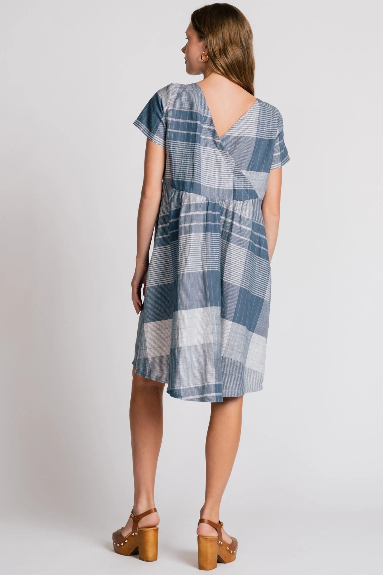 Back view of a  woman wearing the Lark Dress by Allison Wonderland in Blue Plaid print, a short sleeved babydoll dress with pockets and a crossover back. She is standing in front of a plain background.