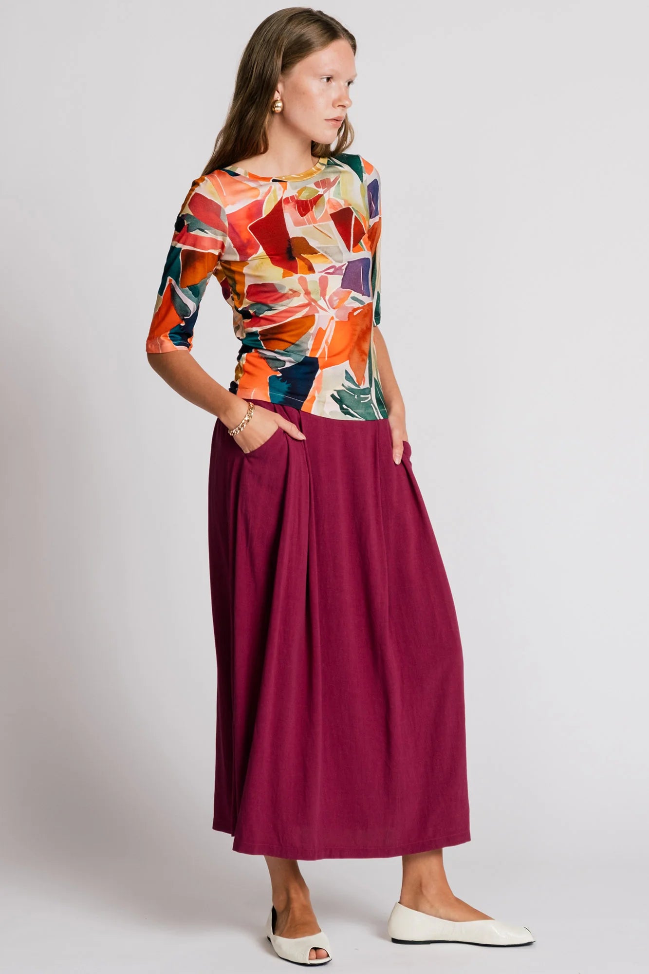 A woman wearing the Lorikeet Top by Allison Wonderland in multicolour print, with a high neck in front, a scooped neck at the back, and 3/4 sleeves. She is wearing it with a burgundy skirt and standing in front of a plain background