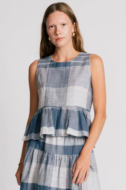 A woman wearing the Osprey Blouse by Allison Wonderland in Blue Plaid, a sleeveless top with a peplum both front and back and a cross-over back. She is wearing it with the matching Pelican Skirt and standing in front of a plain background.  