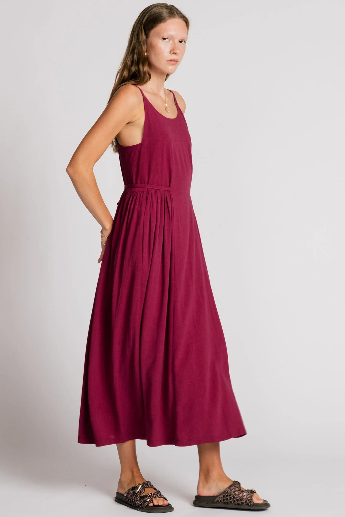 Side view of a woman wearing the Swan Dress by Allison Wonderland in Pink, a maxi dress with spaghetti straps, and ties at the waist. 