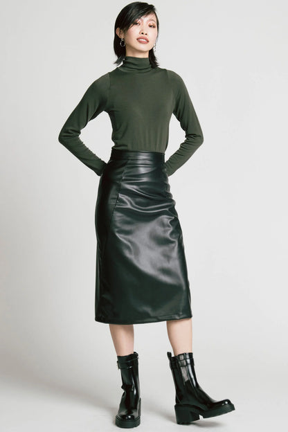 A woman wearing the Desire Skirt by Allison Wonderland in Black faux leather, a midi-length pencil skirt with a two-way back zipper. She is wearing it with a green turtleneck and standing in front of a white background. 