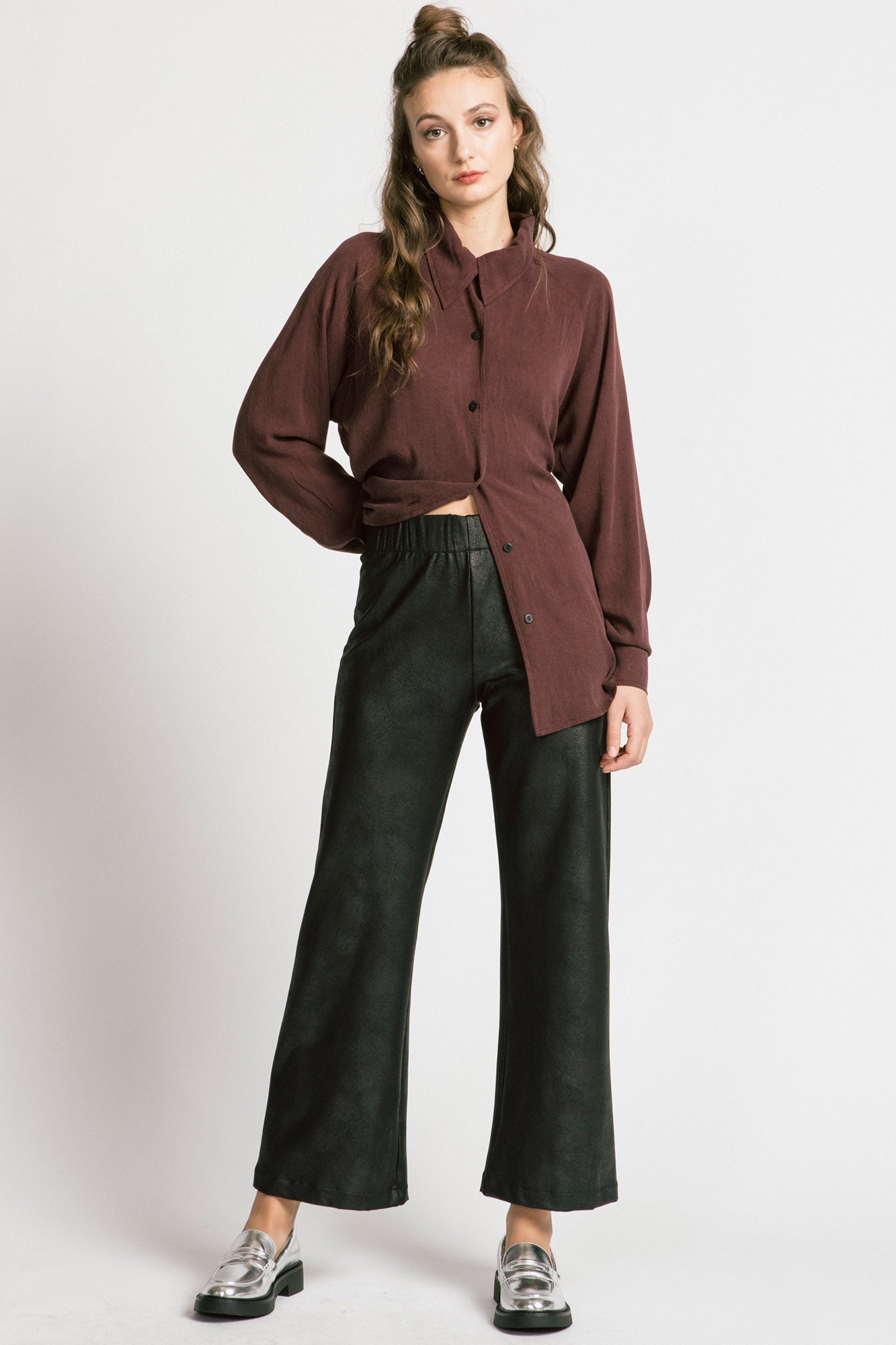 View of a woman wearing the Shanna Pants by Allison Wonderland in Black Leatherette, loose fitting flared pants with an elastic waistband. She is wearing them with a brown top and standing in front of a silver background. 