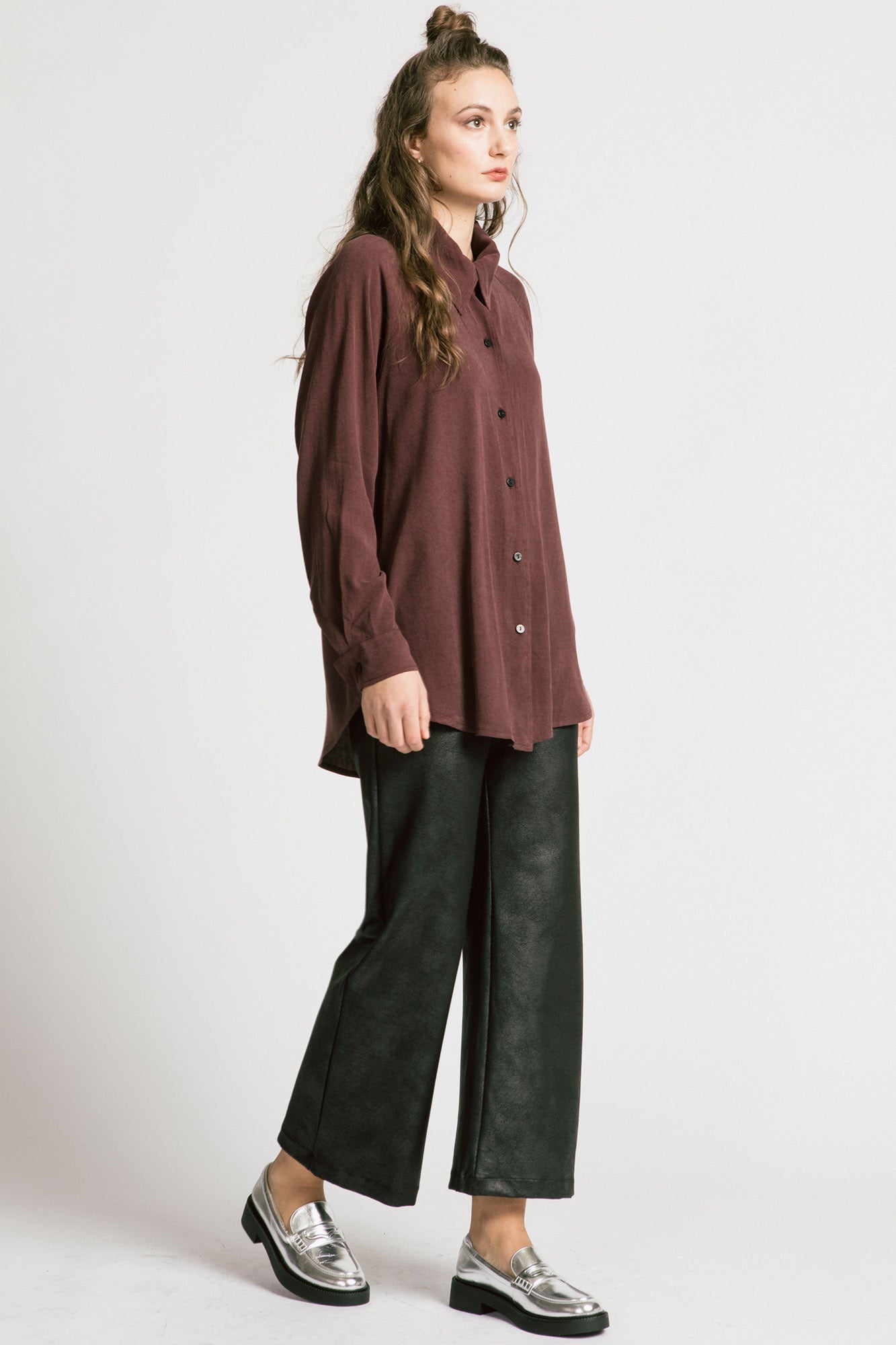 A woman wearing the Shanna Pants by Allison Wonderland in Black Leatherette, loose fitting flared pants with an elastic waistband. She is wearing them with a brown top and standing in front of a silver background. 