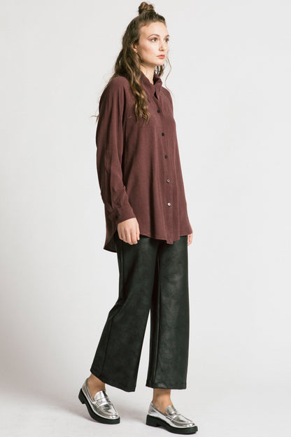 A woman wearing the Shanna Pants by Allison Wonderland in Black Leatherette, loose fitting flared pants with an elastic waistband. She is wearing them with a brown top and standing in front of a silver background. 