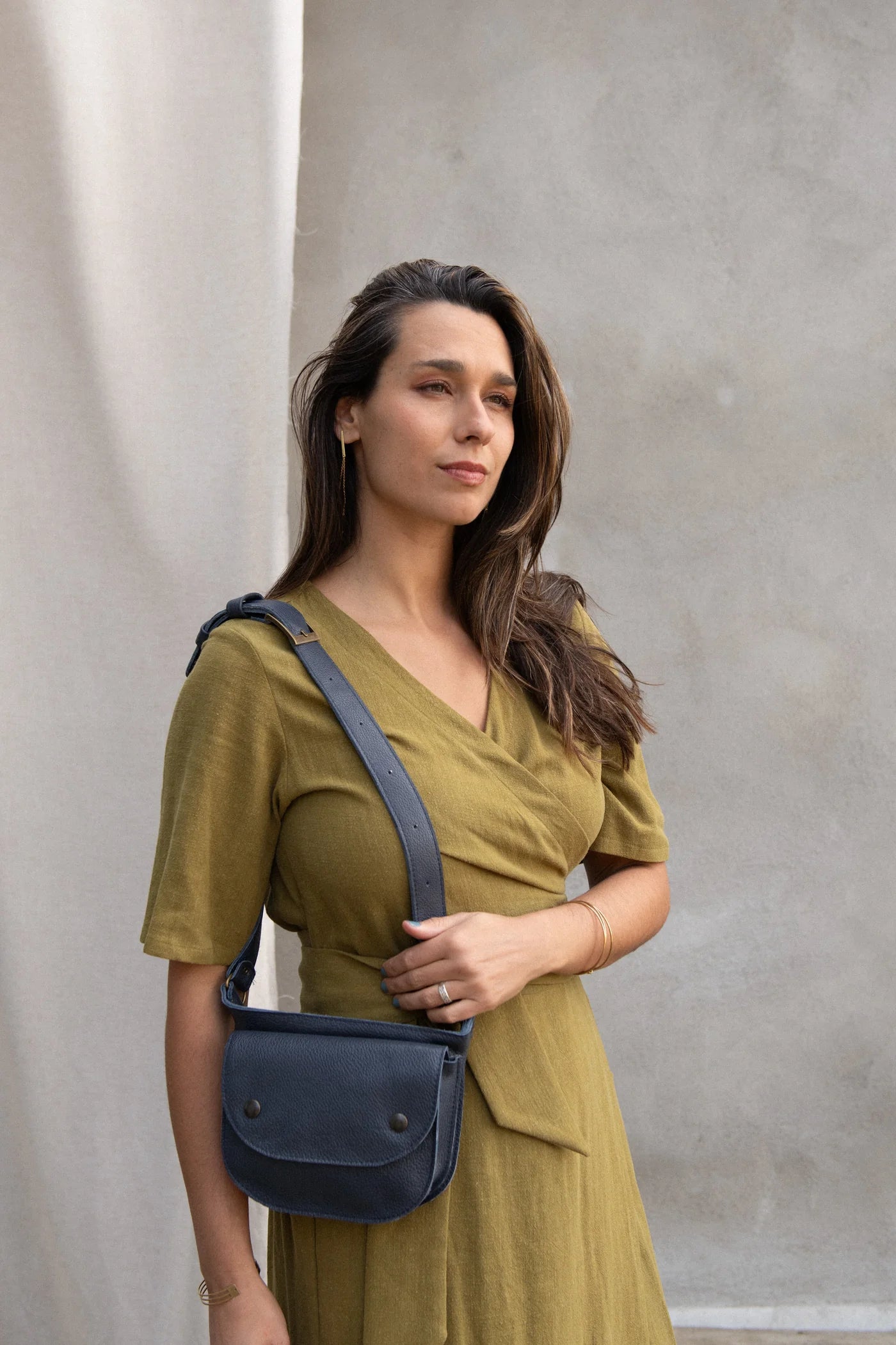 A woman wearing the Helleborus wrap Dress by Kazak in Mousse, standing outside by a white curtain and a cement wall. 