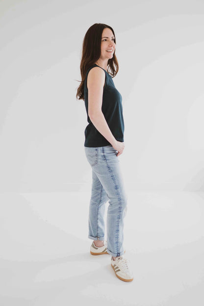 Side view of a woman wearing the Riviera Tank by Ang Hill in Juniper, a loose fitting tank with a round neck and white straps in cotton/modal. She is wearing it with jeans and is standing in front of a white background. 
