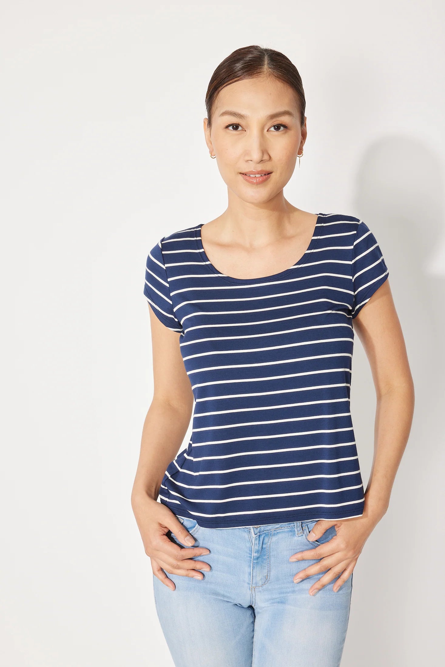 A woman wearing the Fjord T-shirt by Canopée in Navy/White stripe, a short-sleeved tee with a wide scooped neck and a slightly loose fit. She is wearing it with jeans and standing in front of a white background. 