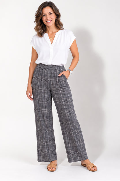 A woman wearing the Sabine pants by Tangente in plaid, linen blend pants with an elastic waist and wide legs in shades of navy/grey/ivory. she is wearing them with a white top and standing in front of a white studio background. 