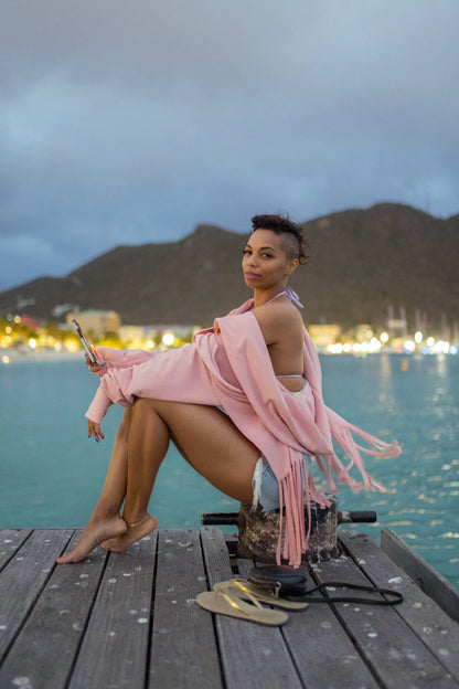 A woman wearing the Heaven Bamboo/Cotton Cropped Wrap from Stacey Martin Lifestyle in Blush Pink, a cropped hoody with a fringed collar scarf that can be tied various ways. She is wearing it with denim shorts and is seated on a dock in front of a lake at twilight. 
