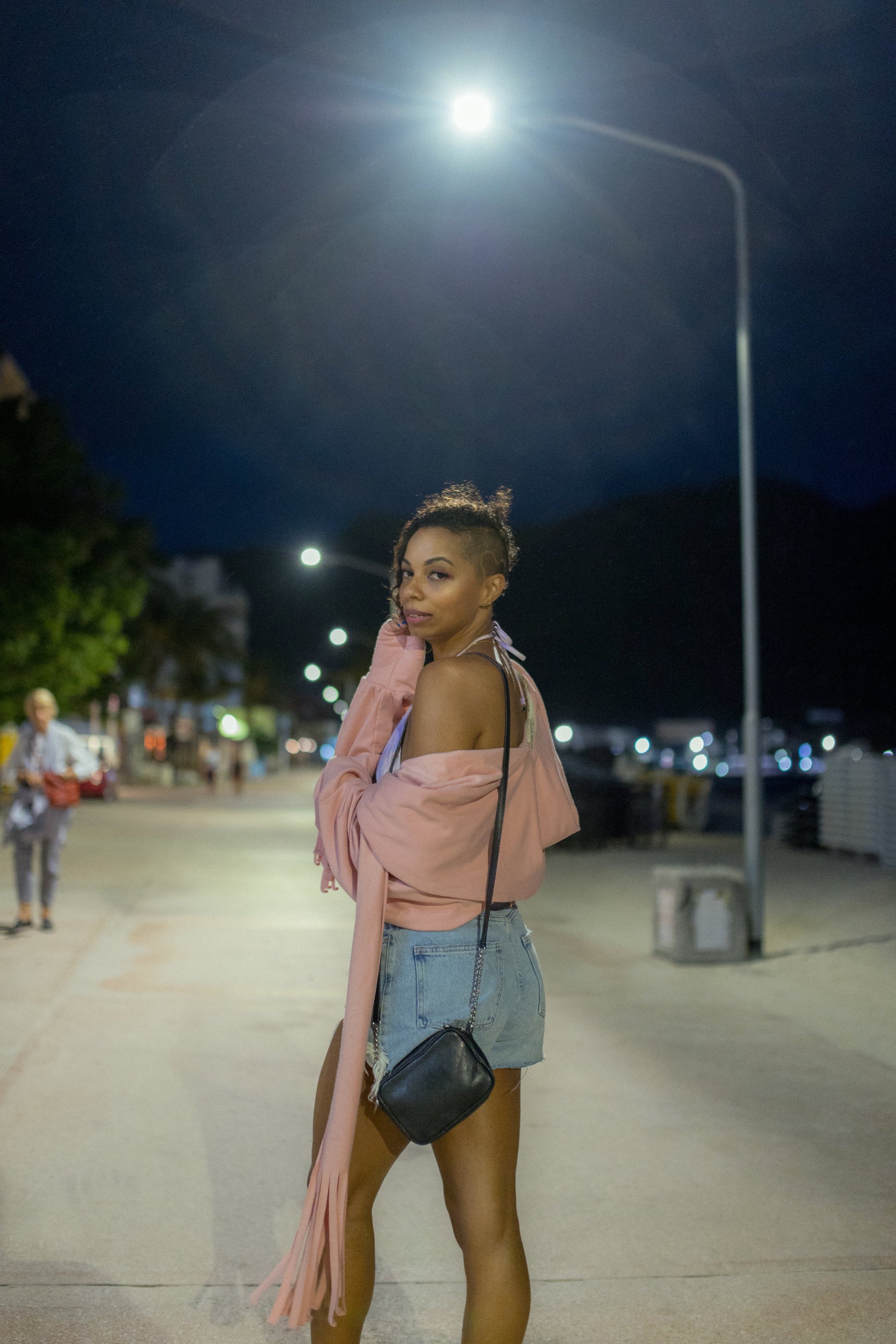A woman wearing the Heaven Bamboo/Cotton Cropped Wrap from Stacey Martin Lifestyle in Blush Pink, a cropped hoody with a fringed collar scarf that can be tied various ways. She is wearing it with denim shorts and is standing on a street at night.