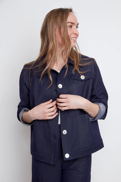 A woman wearing the Bella Knit Jacket by Julei, a denim style jacket with large contrast buttons, a chest pocket and two front patch pockets. She is wearing it with a white and the matching Brooke Knit Trousers and standing in front of a white wall. 