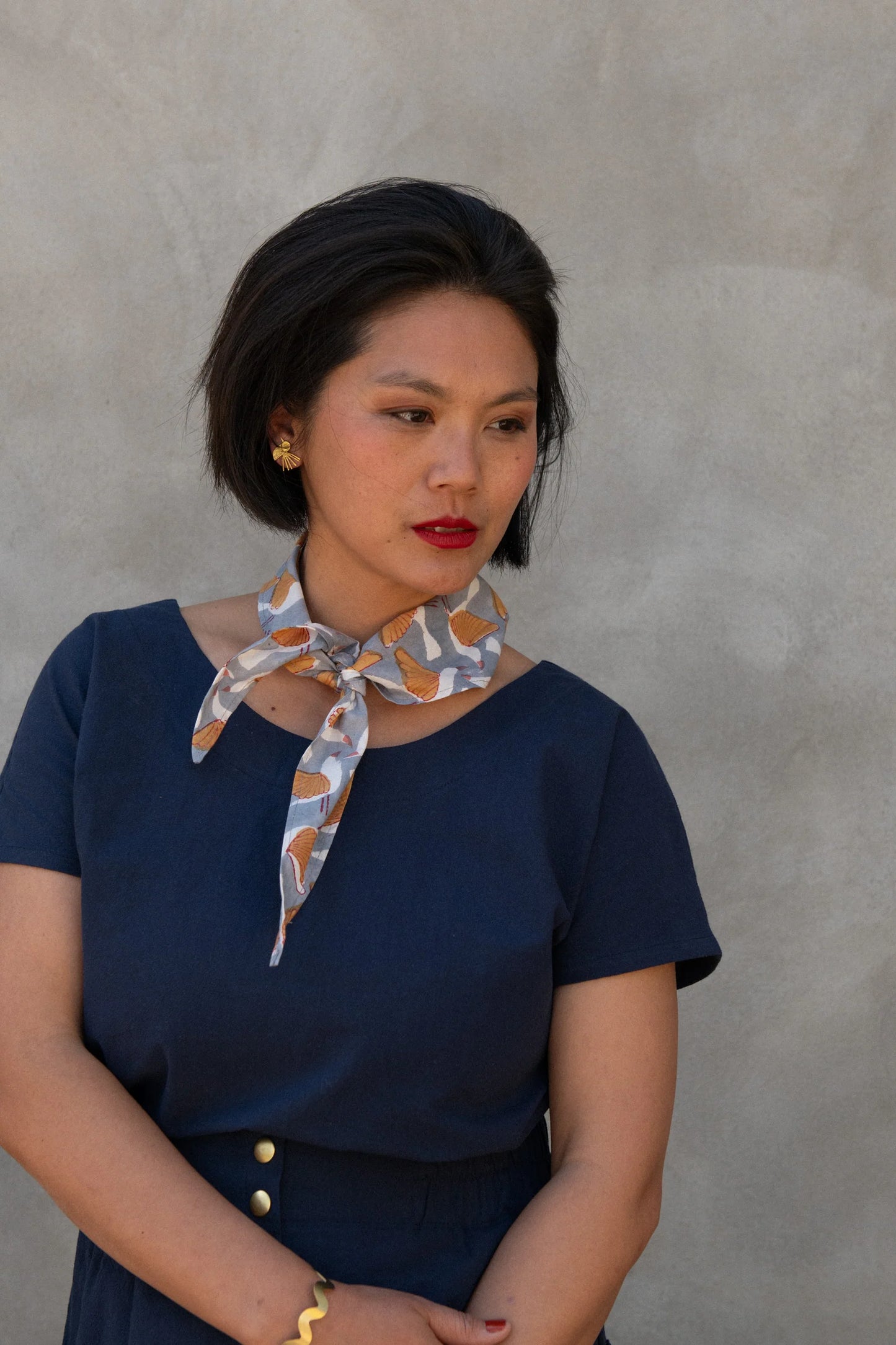 A woman wearing the Brendola Top from Kazak in Navy, a slightly cropped short sleeved top with three brass snaps that can be worn at the back or the front. She is wearing it with a scarf and the Nymphea skirt in Navy and standing in front of a grey wall. 