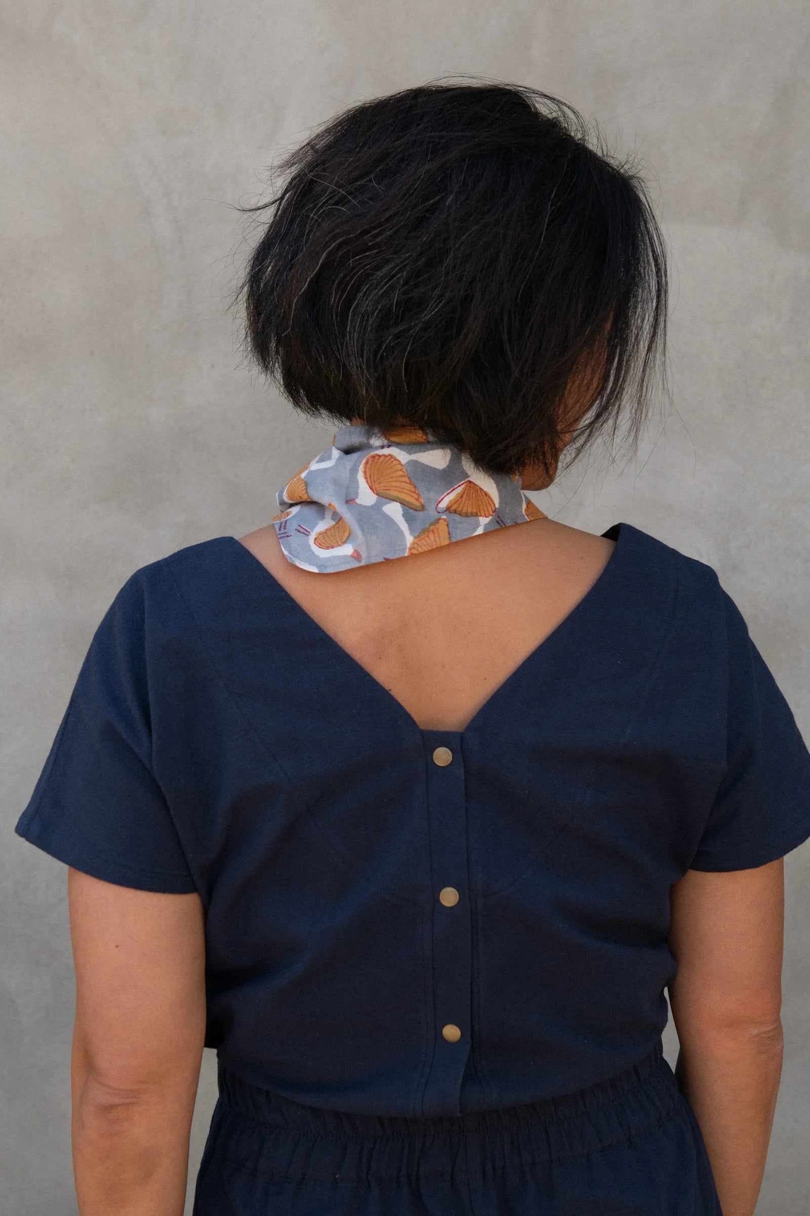Back view of a woman wearing the Brendola Top from Kazak in Navy, a slightly cropped short sleeved top with three brass snaps that can be worn at the back or the front. She is wearing it with a scarf and the Nymphea skirt in Navy and standing in front of a grey wall. 