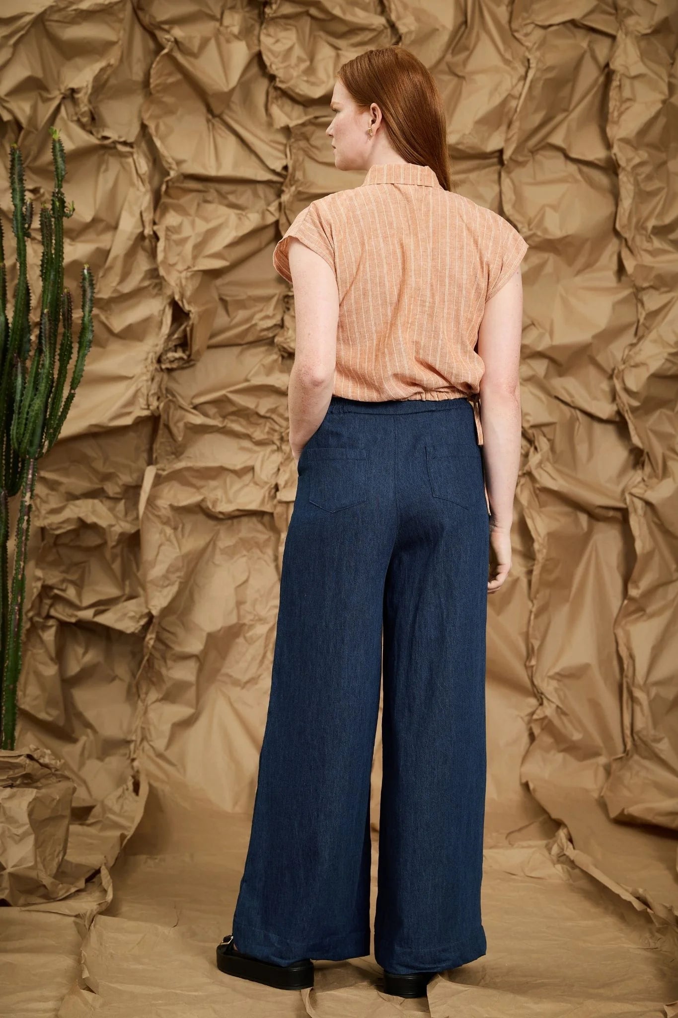 Back view of a woman wearing the Matisse Trousers by Melow, full length wide leg pants with a zipper front and a high waist with elastic at the back with side and back patch pockets. She is wearing them with a peach top and standing in front of a textured brown backdrop