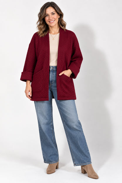 A woman wearing the Ina Coatigan from Tangente in Cherry, a quilted open cardigan with large patch pockets. She is wearing it with a white t-shirt and jeans and standing in front of a white background. 