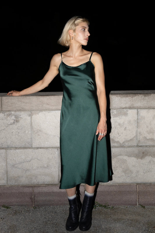 A woman wearing the Celestite Dress by Eve Lavoie in Green Satin, a slip dress with slanted spaghetti straps and an opening at the back with a tie detail. It falls to mid calf. She is standing outdoors by a stone wall. 