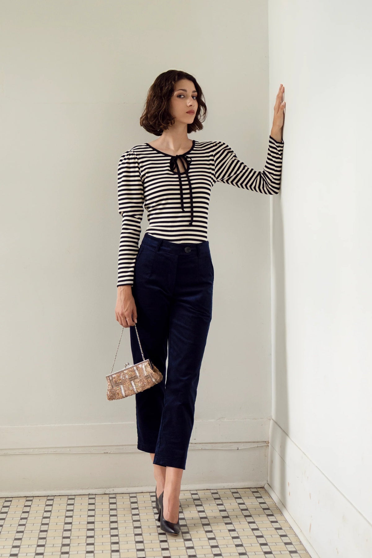 A woman wearing the Cornaline Top by Eve Lavoie, a reversible black and white striped long-sleeve top with a cut-out and tie neckline on one side and a round neckline on the other, plus puff sleeves. She is wearing it with navy cropped pants and standing in a white room. 