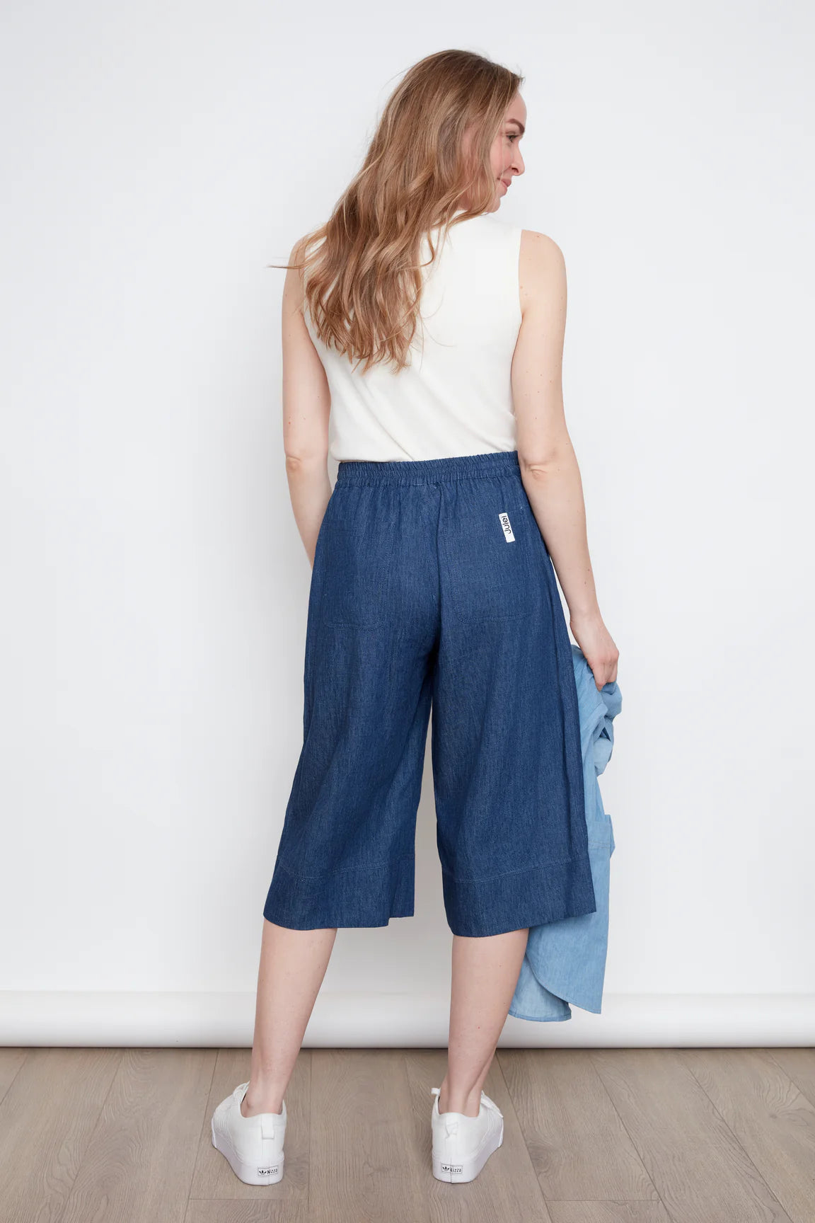 Back view of a woman wearing the Brooke Capri by Julei, wide-legged denim capris with a white drawstring and elastic at the back waist. She is wearing them with a white tank and standing against a white wall.