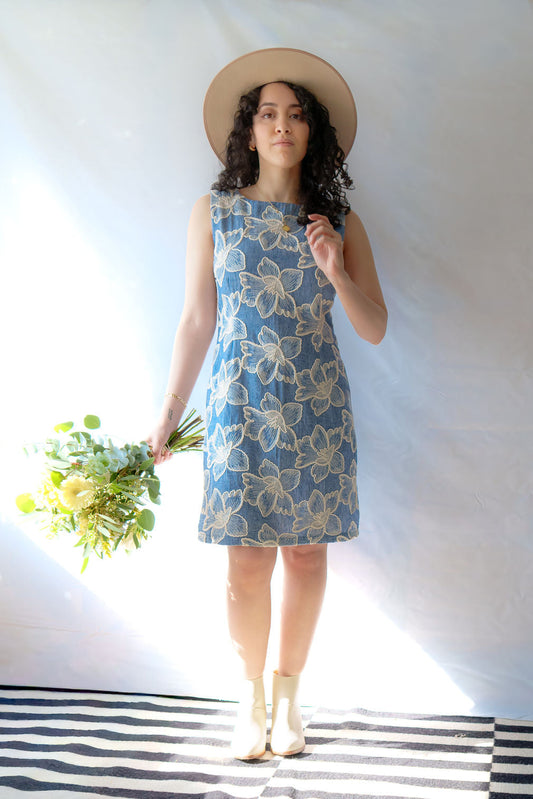 A woman wearing the Rose Dress by Dinh Ba, a sheath dress in pale blue with embroidered white flowers.  She is holding flowers and a tan hat and is  standing in front of a white background