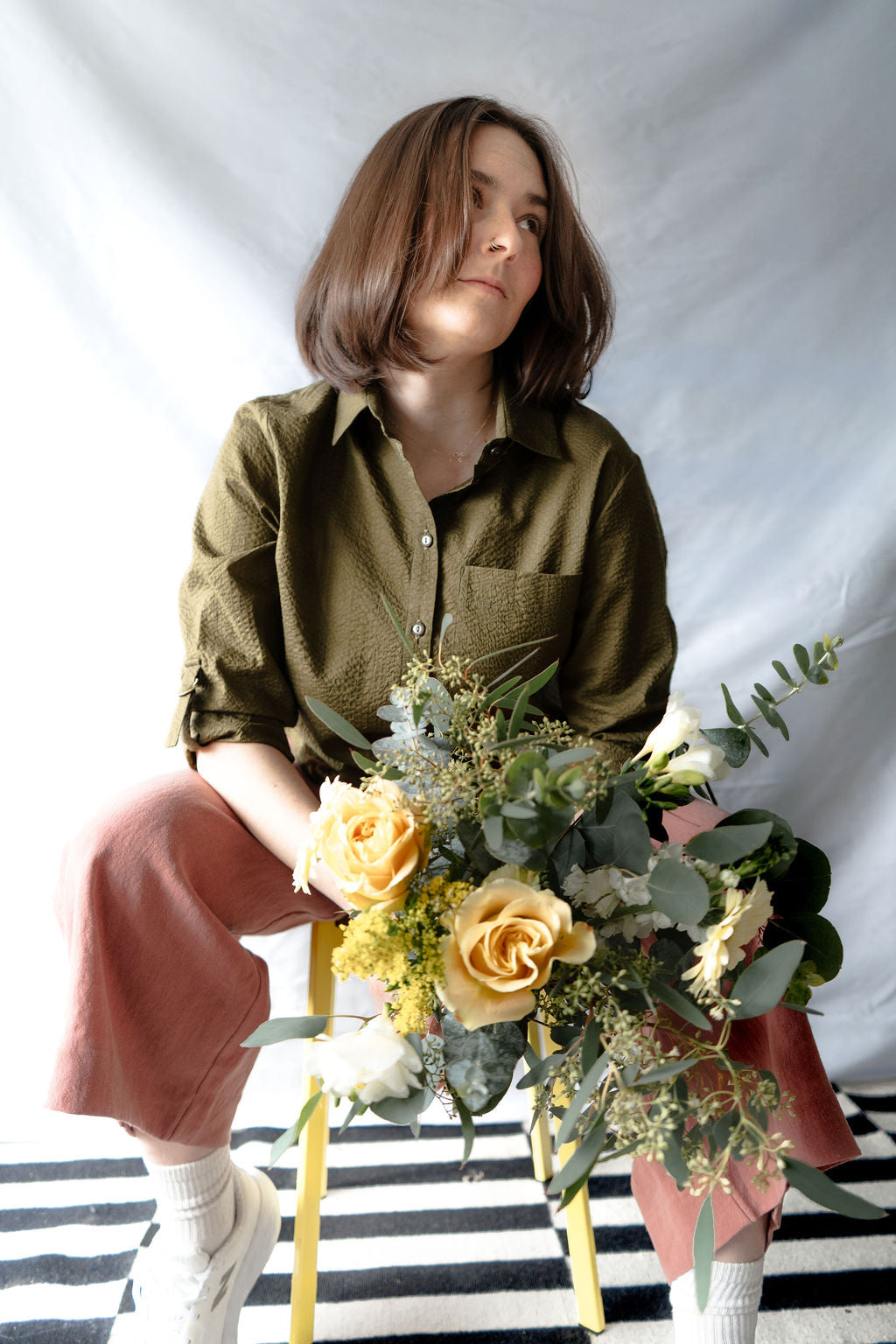 Woman wearing the Ania Blouse by Cokluch in Olive, sitting on a stool and holding a bouquet of flowers on a striped floor with a white background