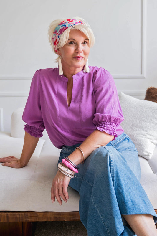 A woman wearing the Orchid Blouse by Dotty in Magenta, with a V-neck with a banded collar and elbow length smocked sleeves. She is wearing it with jeans and sitting on a couch.