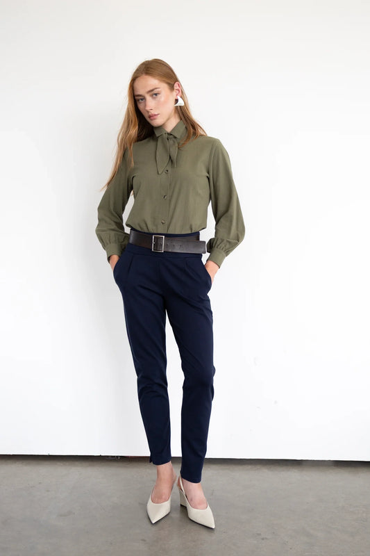A woman wearing the Fraser Pants by Eve Lavoie in Navy, Ponte di Roma pants with a wide pull-on waistband, slanted side pockets, and an ankle-length leg with a slit detail. She is wearing them with a green top and standing in front of a white wall. 