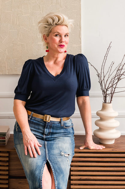 A woman wearing the FW Darling Tee by Dotty in Navy, featuring a v-neck, pleated shoulders, and elbow length sleeves, with a jean skirt. She is sitting in a room with a vase and branches on a table.