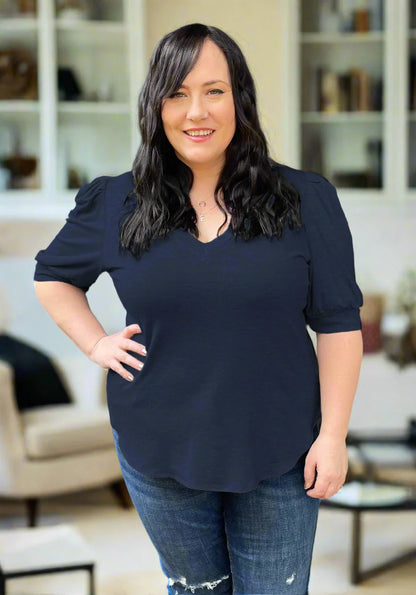 A woman wearing the FW Darling Tee by Dotty in Navy, featuring a v-neck, pleated shoulders, and elbow length sleeves, with jeans. She is standing indoors in front of a couch.