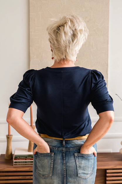 Back view of a woman wearing the FW Darling Tee by Dotty in Navy, featuring a v-neck, pleated shoulders, and elbow length sleeves, with a jean skirt. She is standing in front of table. 