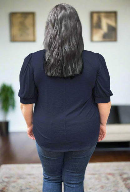 Back view of a woman wearing the FW Darling Tee by Dotty in Navy, featuring a v-neck, pleated shoulders, and elbow length sleeves, with a jeans. She is standing in front of a couch.