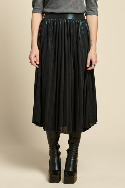 Waist down view of a woman wearing the Feyer Skirt by Cokluch in Glow Knit Black, a midi length pleated skirt in shimmery metallic fabric. She is wearing it with the Insomie top in Facet Black and is standing in front of a beige background. 