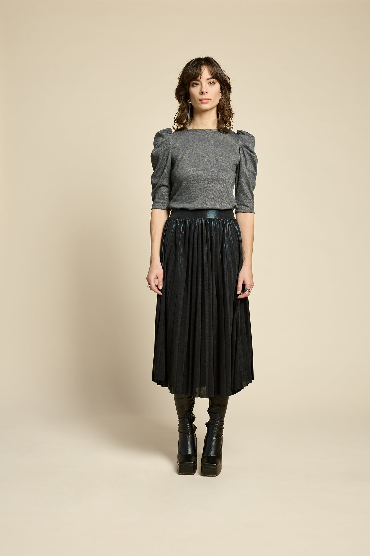 A woman wearing the Feyer Skirt by Cokluch in Glow Knit Black, a midi length pleated skirt in shimmery metallic fabric. She is wearing it with the Insomie top in Facet Black and is standing in front of a beige background. 