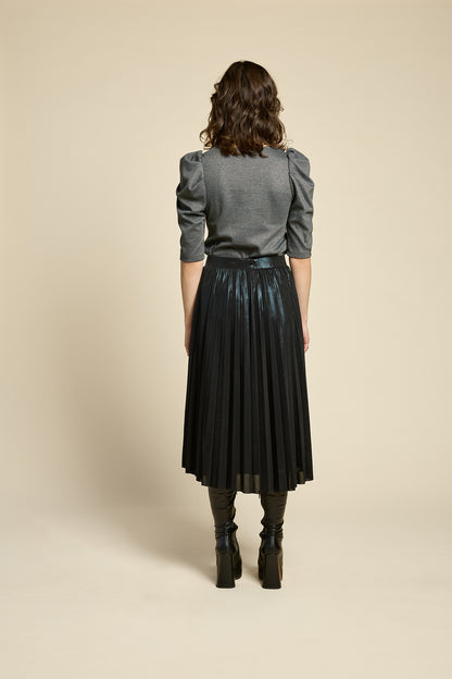 Back view of a woman wearing the Feyer Skirt by Cokluch in Glow Knit Black, a midi length pleated skirt in shimmery metallic fabric. She is wearing it with the Insomie top in Facet Black and is standing in front of a beige background. 