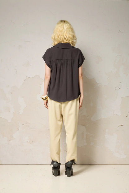 Back view of a woman wearing the Hemera Shirt by Cokluch in Iron, with a V-neck collar, button front, short extended sleeves, and a relaxed fit. The hem is hip-length in the front and longer at the back. She's wearing it with beige pants and standing in front of a grey background. 
