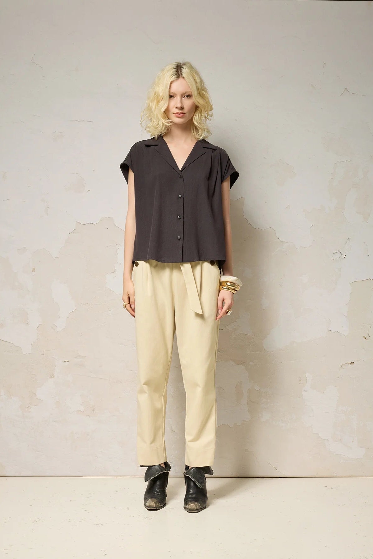 A woman wearing the Hemera Shirt by Cokluch in Iron, with a V-neck collar, button front, short extended sleeves, and a relaxed fit. The hem is hip-length in the front and longer at the back. She's wearing it with beige pants and standing in front of a grey background. 