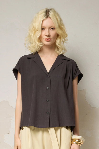 Waist up view of a woman wearing the Hemera Shirt by Cokluch in Iron, with a V-neck collar, button front, short extended sleeves, and a relaxed fit. The hem is hip-length in the front and longer at the back. She's wearing it with beige pants and standing in front of a grey background. 