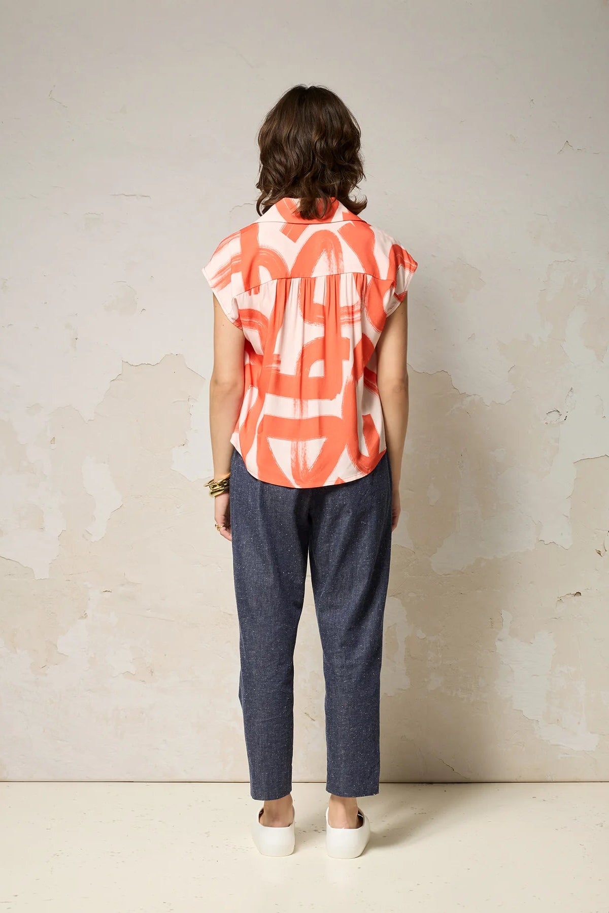 Back view of a woman wearing the Hemera Shirt by Cokluch in Red Shodo, with a V-neck collar, button front, short extended sleeves, and a relaxed fit. The hem is hip-length in the front and longer at the back. She's wearing it with denim pants and standing in front of a grey background. 