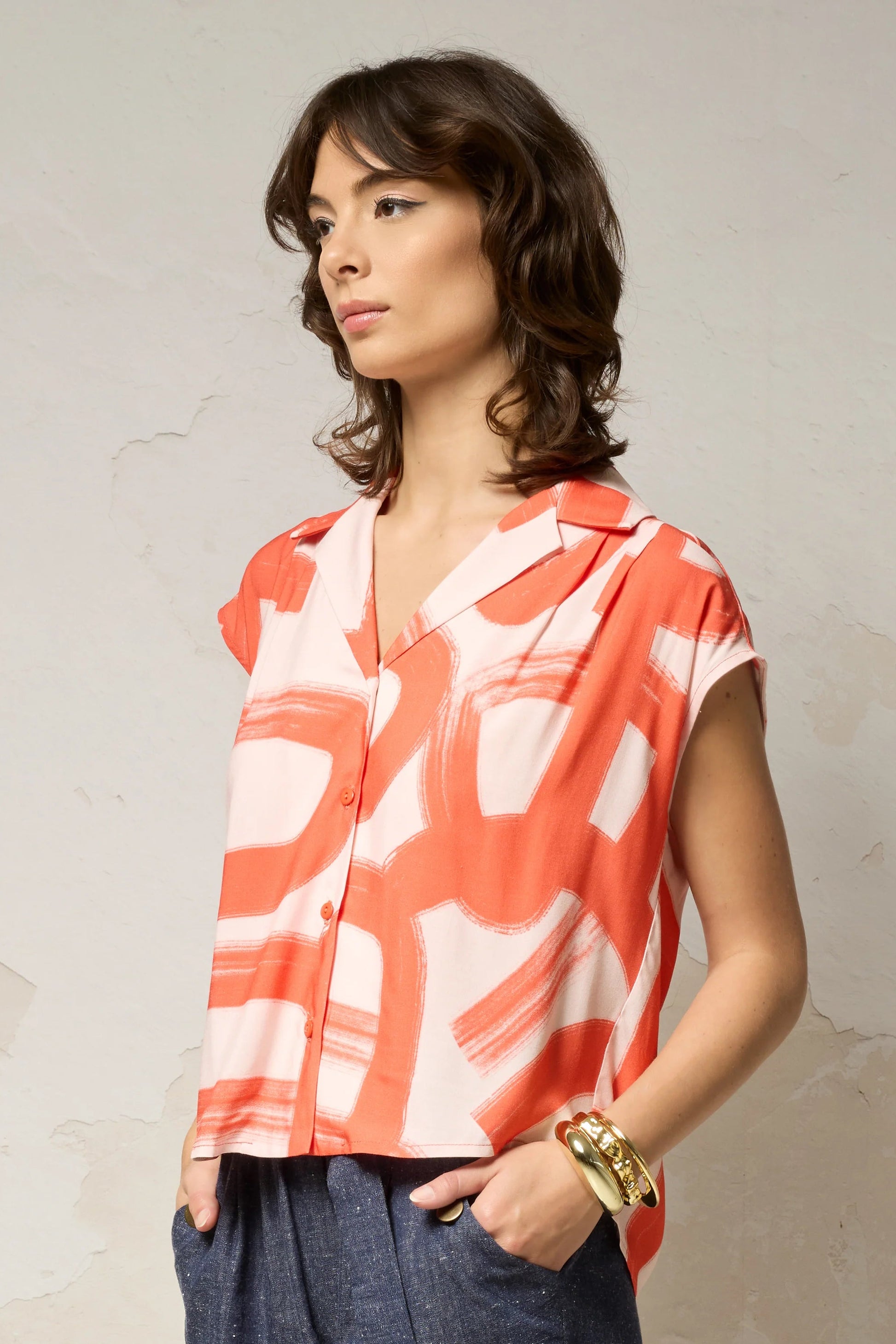 Waist up view of a woman wearing the Hemera Shirt by Cokluch in Red Shodo print, with a V-neck collar, button front, short extended sleeves, and a relaxed fit. The hem is hip-length in the front and longer at the back. She's wearing it with denim pants and standing in front of a grey background. 
