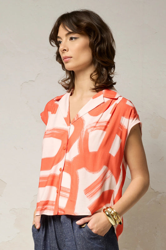 Waist up view of a woman wearing the Hemera Shirt by Cokluch in Red Shodo print, with a V-neck collar, button front, short extended sleeves, and a relaxed fit. The hem is hip-length in the front and longer at the back. She's wearing it with denim pants and standing in front of a grey background. 
