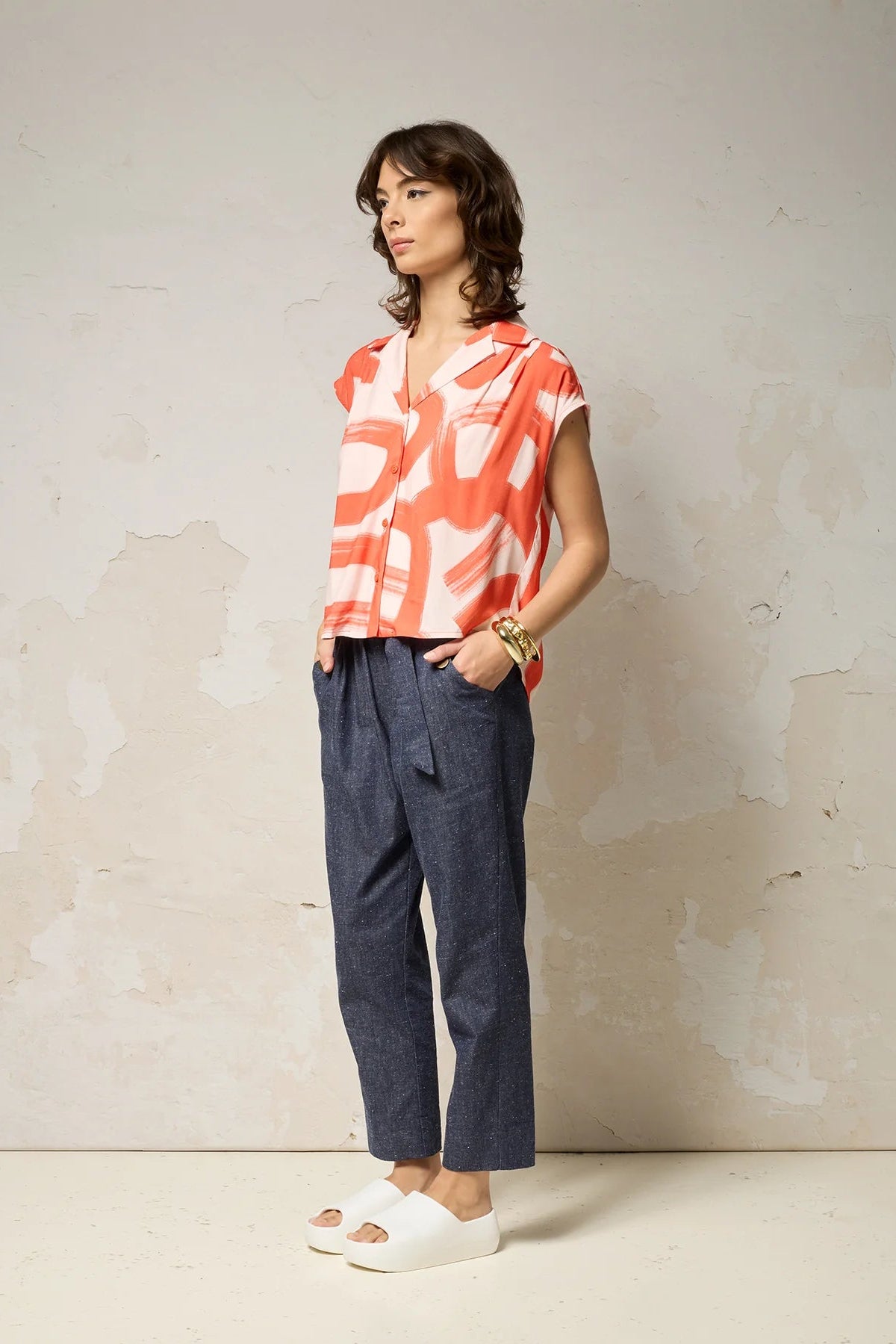 A woman wearing the Hemera Shirt by Cokluch in Red Shodo, with a V-neck collar, button front, short extended sleeves, and a relaxed fit. The hem is hip-length in the front and longer at the back. She's wearing it with denim pants and standing in front of a grey background. 