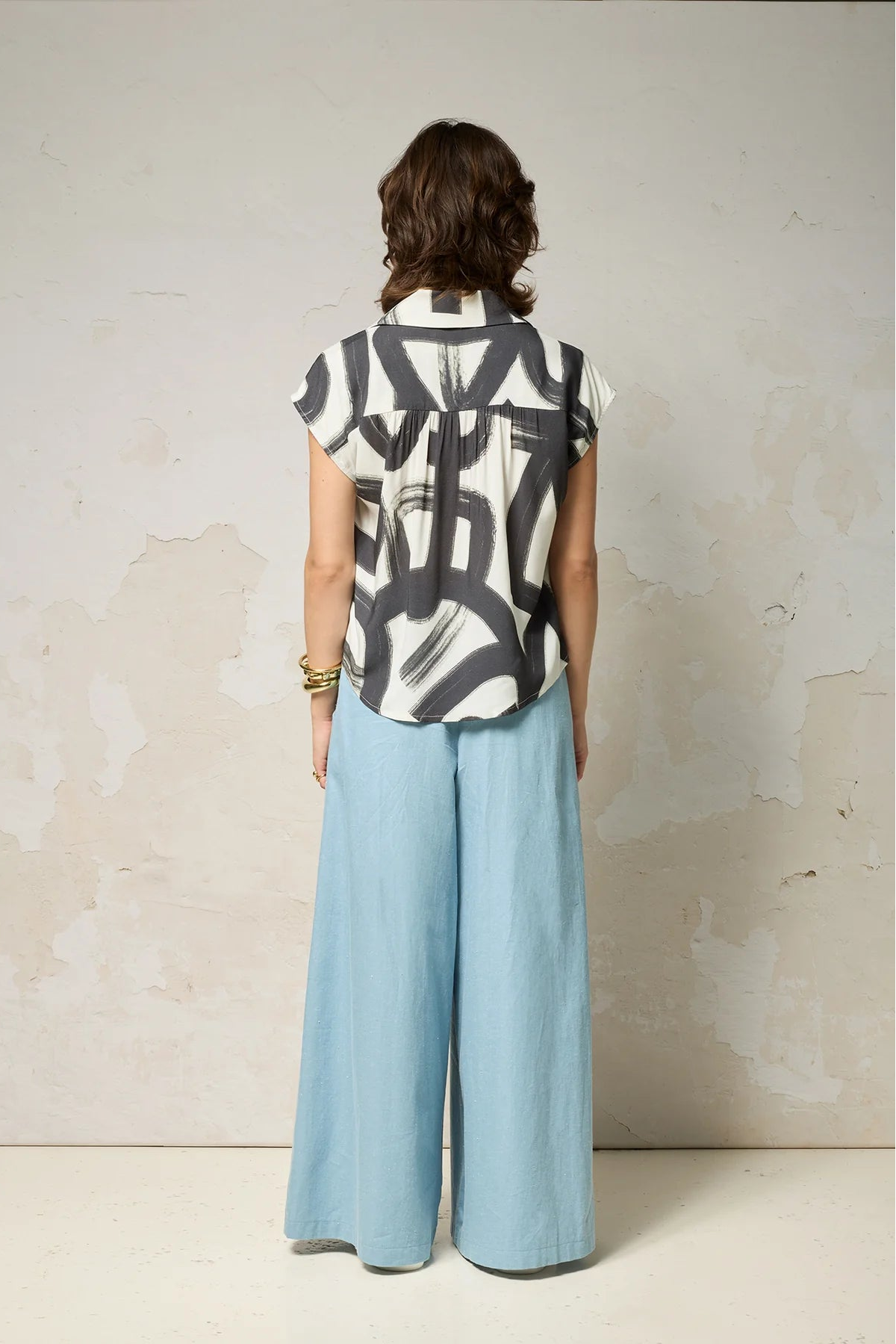 Back view of a woman wearing the Hemera Shirt by Cokluch in Black Shodo, with a V-neck collar, button front, short extended sleeves, and a relaxed fit. The hem is hip-length in the front and longer at the back. She's wearing it with wide legged blue pants and standing in front of a grey background. 