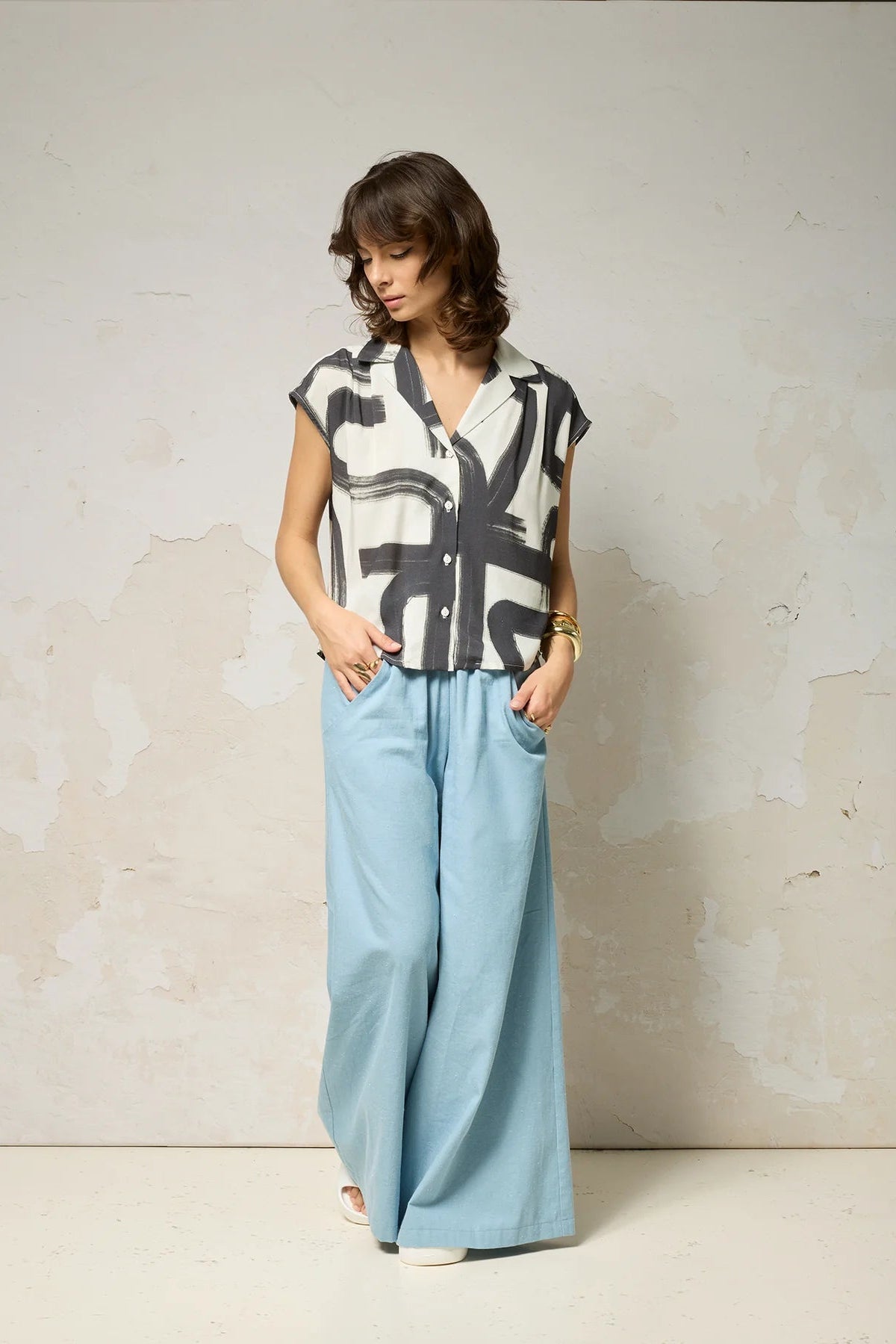 A woman wearing the Hemera Shirt by Cokluch in Black Shodo, with a V-neck collar, button front, short extended sleeves, and a relaxed fit. The hem is hip-length in the front and longer at the back. She's wearing it with wide legged blue pants and standing in front of a grey background. 