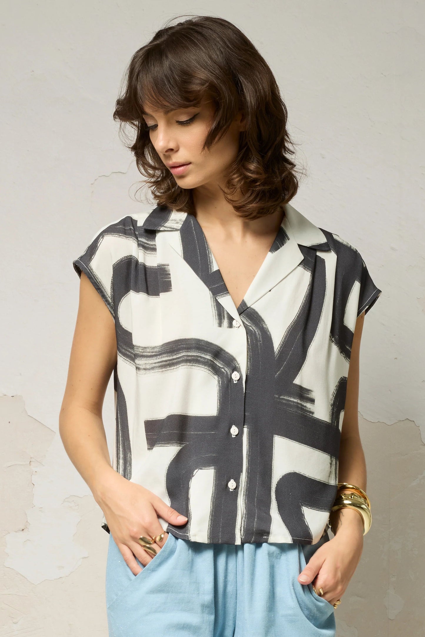 Waist up view of a woman wearing the Hemera Shirt by Cokluch in Black Shodo print, with a V-neck collar, button front, short extended sleeves, and a relaxed fit. The hem is hip-length in the front and longer at the back. She's wearing it with blue pants and standing in front of a grey background. 
