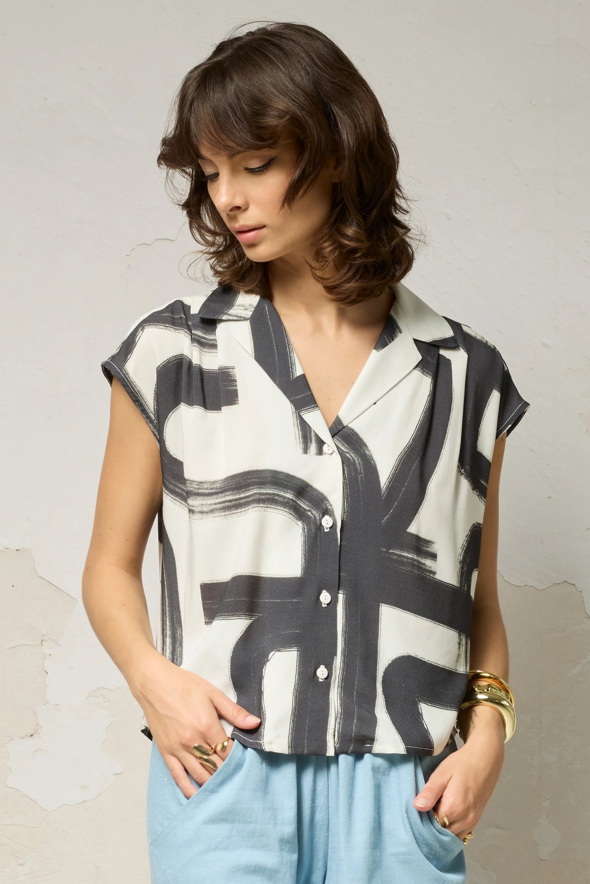 Waist up view of a woman wearing the Hemera Shirt by Cokluch in Black Shodo print, with a V-neck collar, button front, short extended sleeves, and a relaxed fit. The hem is hip-length in the front and longer at the back. She's wearing it with blue pants and standing in front of a grey background. 