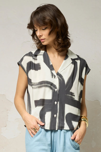 Waist up view of a woman wearing the Hemera Shirt by Cokluch in Black Shodo print, with a V-neck collar, button front, short extended sleeves, and a relaxed fit. The hem is hip-length in the front and longer at the back. She's wearing it with blue pants and standing in front of a grey background. 