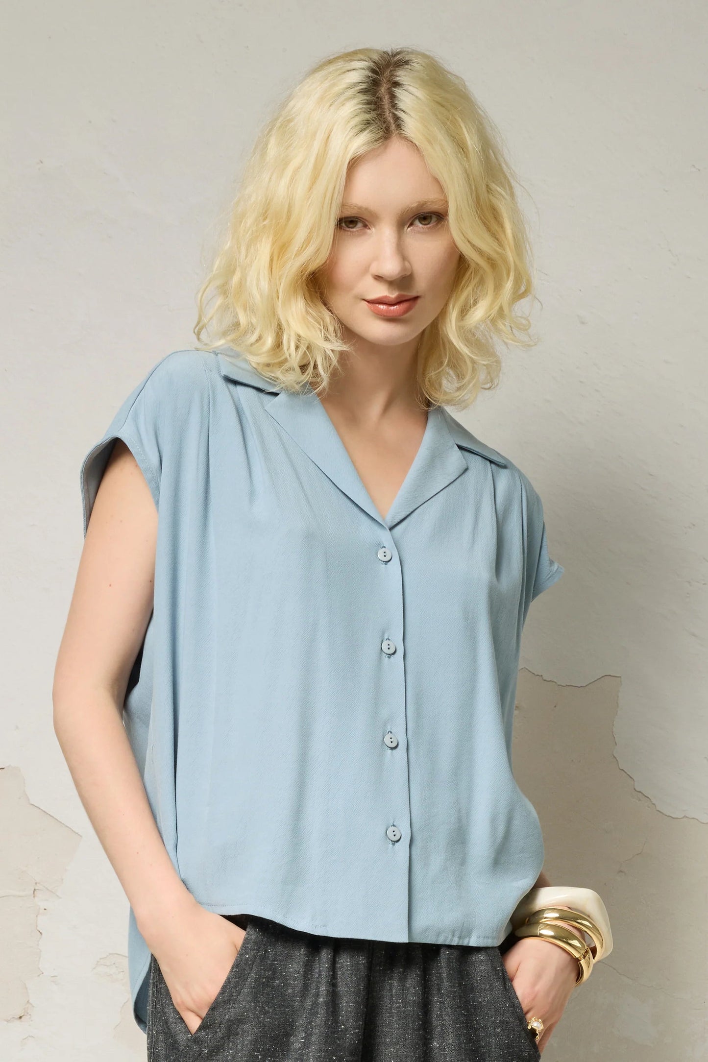 Waist up view of a woman wearing the Hemera Shirt by Cokluch in Sky, with a V-neck collar, button front, short extended sleeves, and a relaxed fit. The hem is hip-length in the front and longer at the back. She's wearing it with denim pants and standing in front of a grey background. 